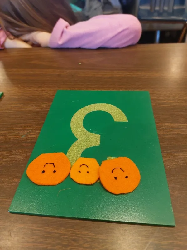 On a table, there is a paper with the number 3 on it with 3 orange smiley face cutouts on it. A child’s arm is resting on the table.