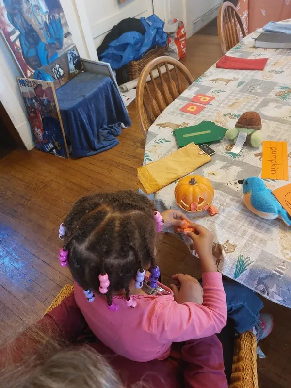 A child sitting on an adult’s lap at a table, putting orange play-dough on a mini jack-o-lantern.