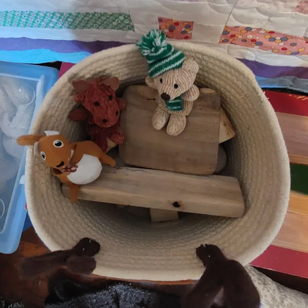 A small plush squirrel, moose, and bear with a winter hat and scarf sit on wooden shapes inside a round bin with fuzzy antlers attached.
