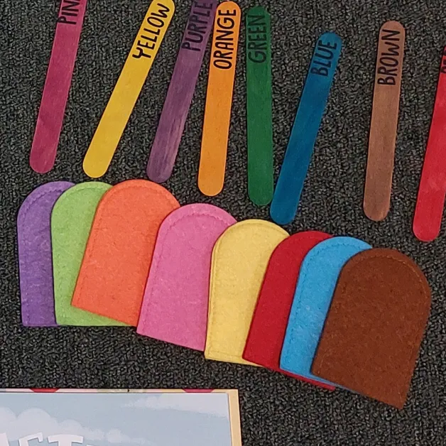 Popsicle sticks with color names and felt popsicles for each color laid out on a rug.