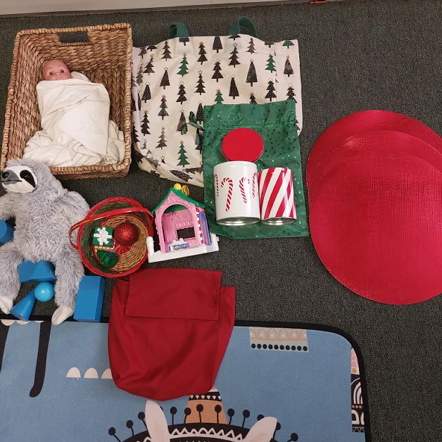 An assortment of objects of different shapes - baby Jesus in a rectangular basket, rectangular and square bags, cylinder-shaped canisters, ornaments in a circle-shaped basket, a triangular/rectangular shaped toy building, and Montessori geometric solids next to a stuffed animal.