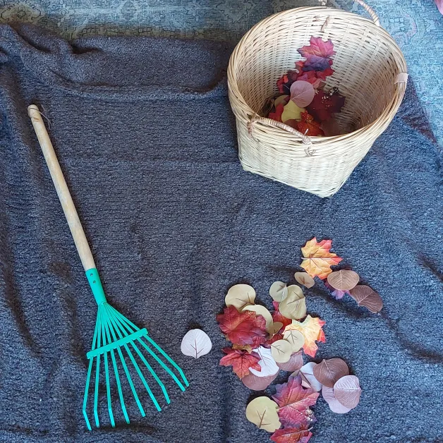 A photo of a kid-sized rake and fall leaves on a blanket. Some of the leaves have been gathered into a basket.