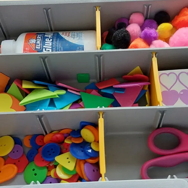 Closeup photo of a sectioned container with craft supplies: a glue bottle, pom-poms, foam shapes, heart stickers, buttons, and a pair of scissors.