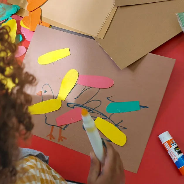 A child creating a thankful turkey - the turkey is created by tracing the child's hand, with a drawn eye, beak, and feet, and paper feather cutouts glued on each finger outline.